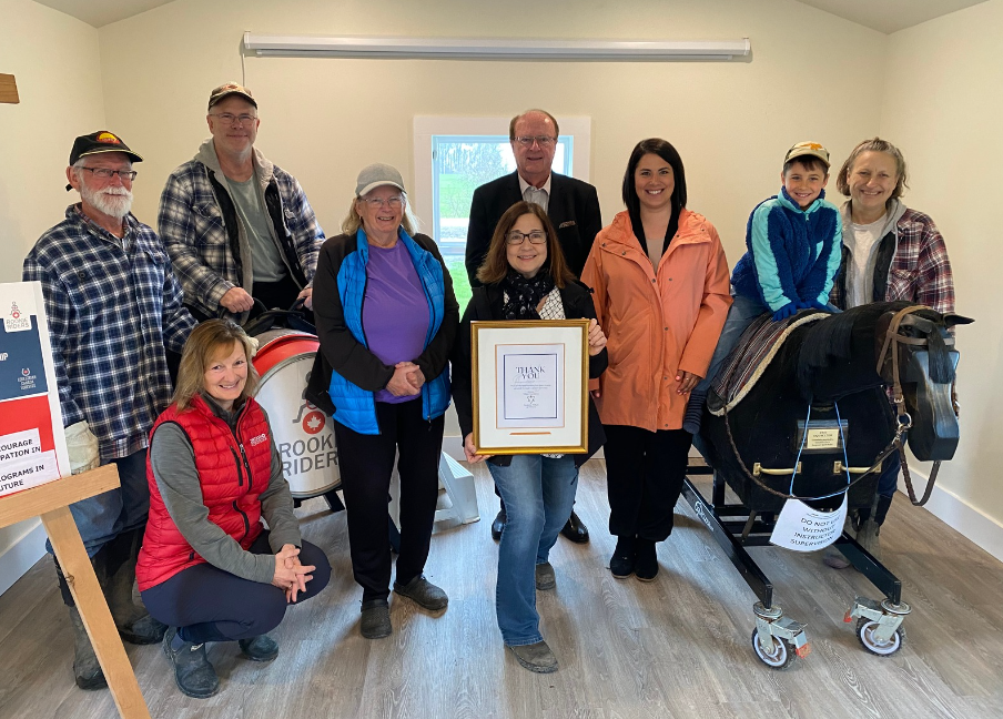 TEAD volunteers, staff, Board Members, and representatives of Donna Skelly smiling at the camera inside the clubhouse. A Board Member is holding a thank you plaque for OTF.