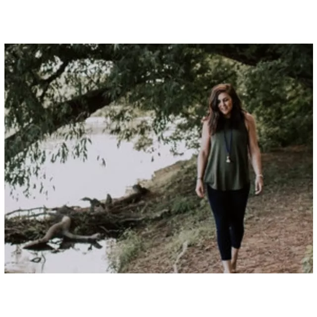 A brown hair female stands next to a river. She is looking down. Green trees are behind her.